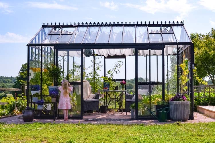 Frontal vy av ett stilrent glasväxthus med svart ram, dekorerat med växter och utemöbler, samt en liten flicka i ljus klänning som tittar in.