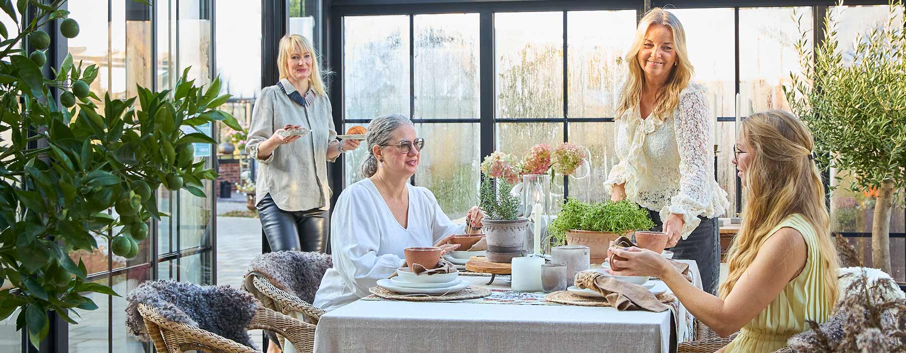 Fyra kvinnor njuter av en mysig samvaro vid ett vackert uppdukat bord i ett CL Alu uterum i viktoriansk stil. Det isolerade uterummet från Classicum är tillverkat i Sverige och har eleganta svarta aluminiumramar med stora glaspartier som släpper in massor av naturligt ljus. Kvinnorna sitter vid ett bord med linneduk, keramikskålar och krukor med gröna växter. Uterummet är inrett med gröna växter och har en varm, inbjudande atmosfär trots den kalla årstiden utanför. Den viktorianska stilen på konstruktionen skapar en tidlös elegans medan den moderna isoleringen gör det möjligt att använda utrymmet året runt.