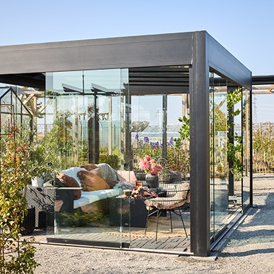 Fristående aluminiumpergola med glasade väggar, inredd som sittplats med möbler och växter, placerad i trädgårdsmiljö. Kan även monteras mot hus.