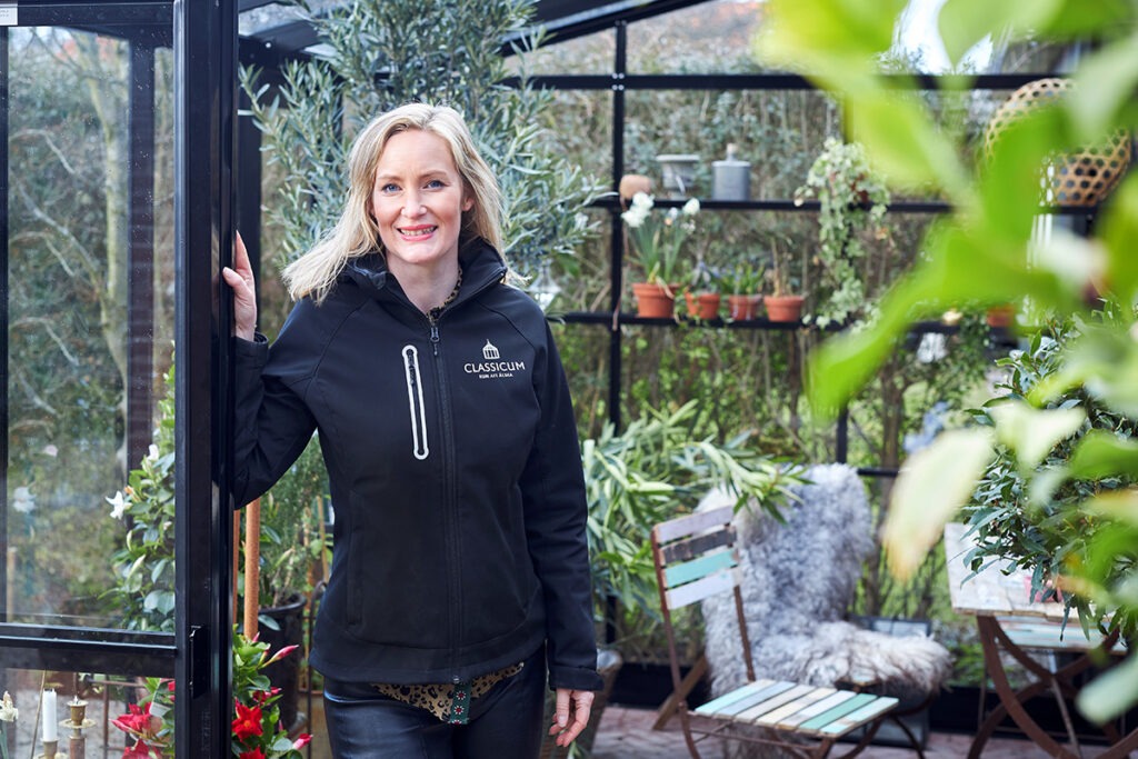 Kvinna står i ett Classicum-växthus omgiven av grönskande växter och inredning.