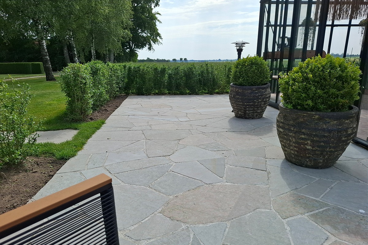 Stenlagd uteplats intill Antique-växthus från Classicum, med planteringskärl och häck som ramar in platsen.