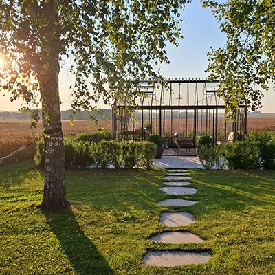 Växthus av modellen Antique från Classicum sett genom träd, med trampstensgång och utsikt över öppna fält.