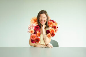 Kvinna med jord på händer och kind, omgiven av färgstarka dahlior runt axlarna, sittande vid ett bord mot ljus bakgrund.