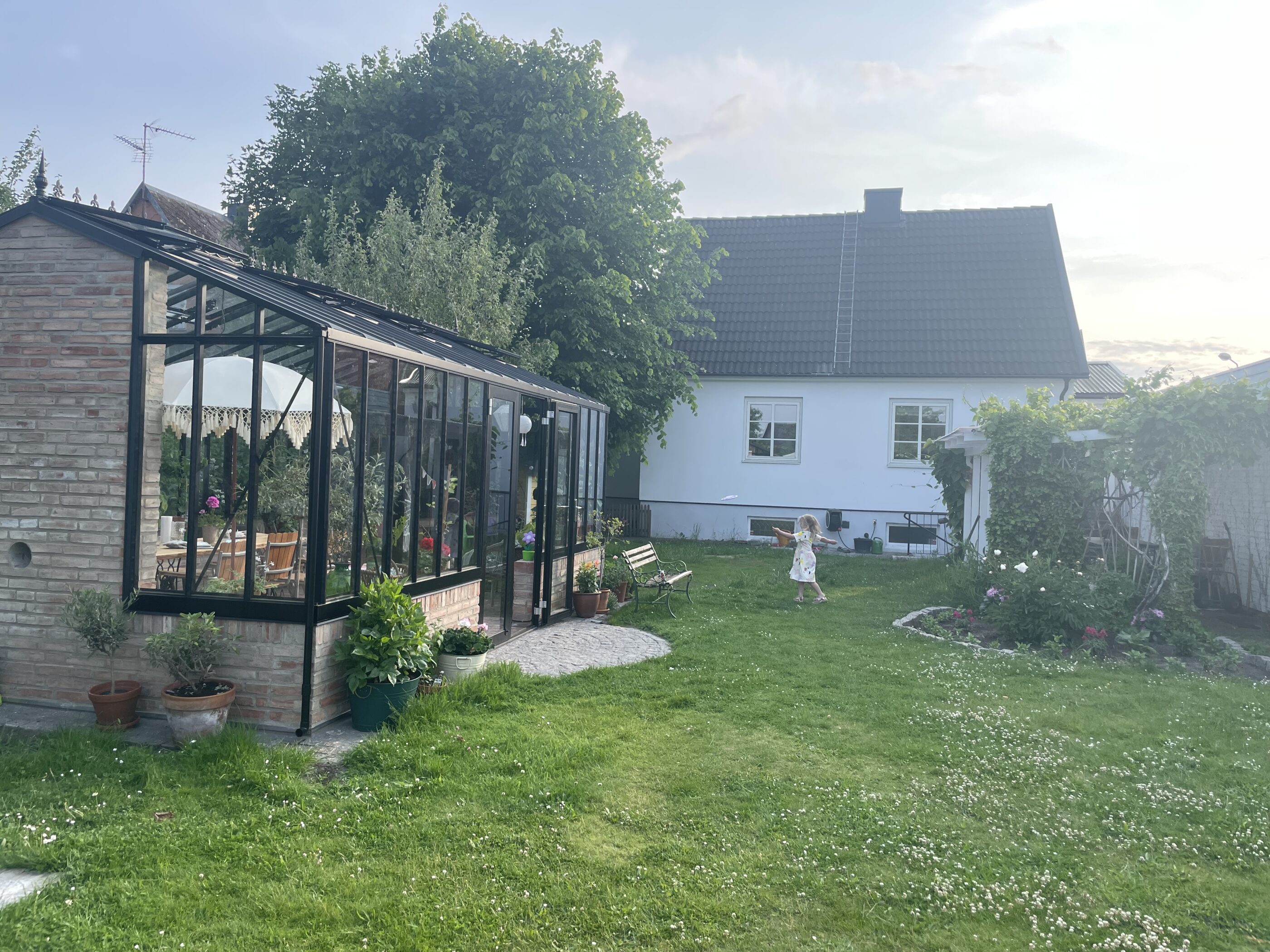 Somrig bild på ett växthus i en trädgård med hus i bakgrunden och ett barn som leker