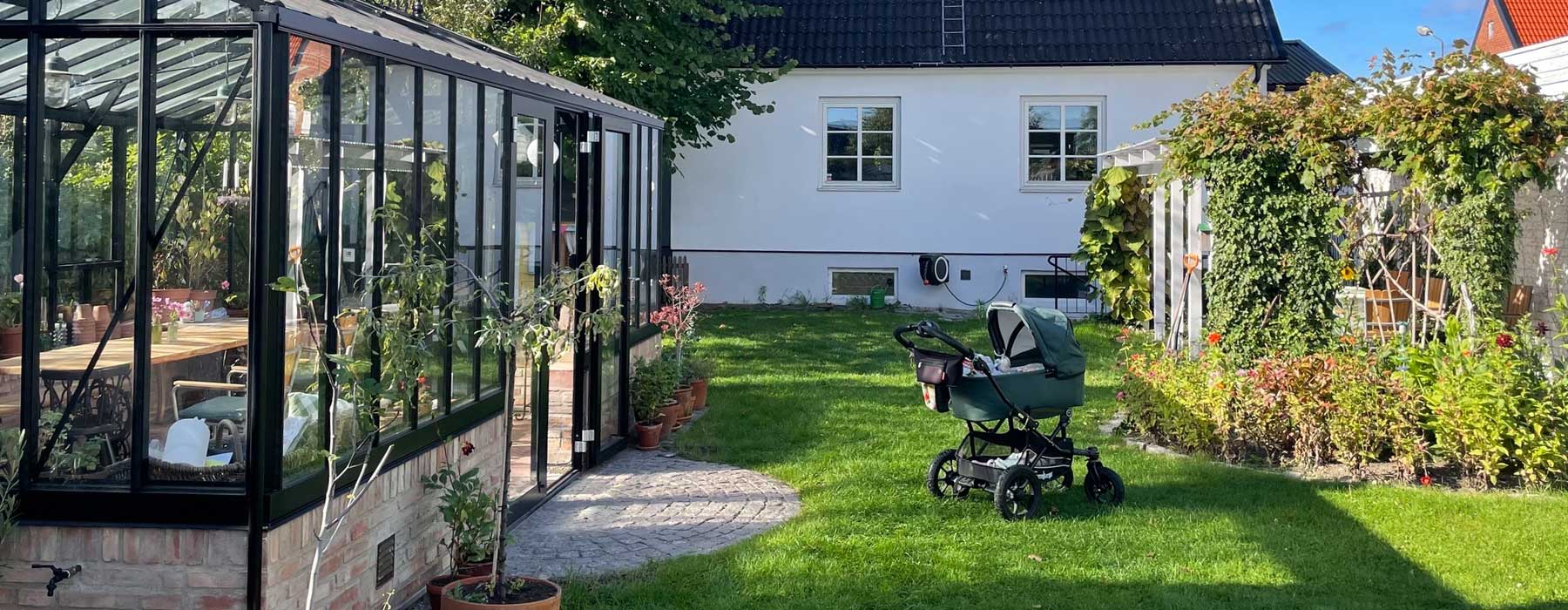 trädgård med växthus, vitt bostadshus och barnvagn på gräset